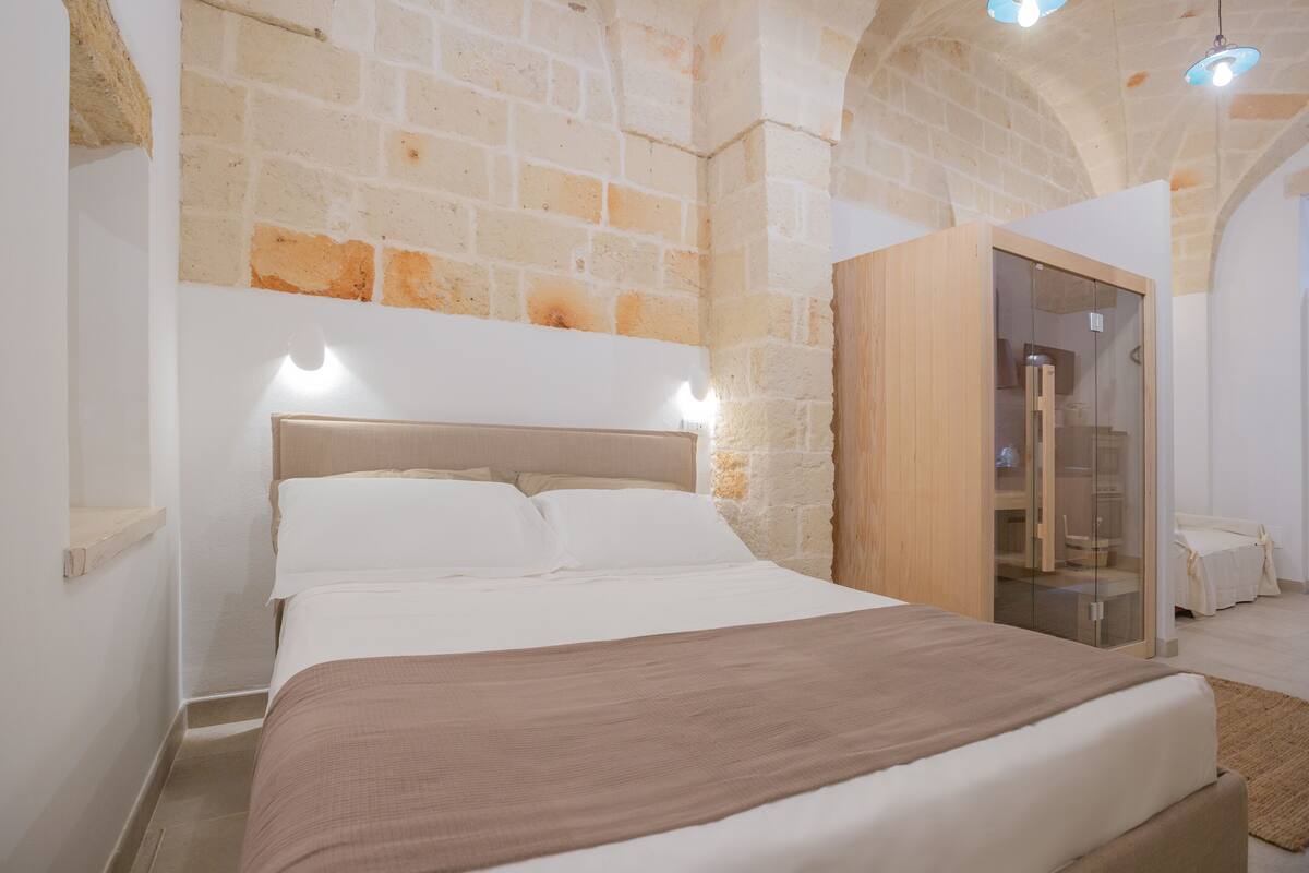 A bedroom with a brick wall, a wooden wardrobe, a bed with white linens, and a small wooden shelf.