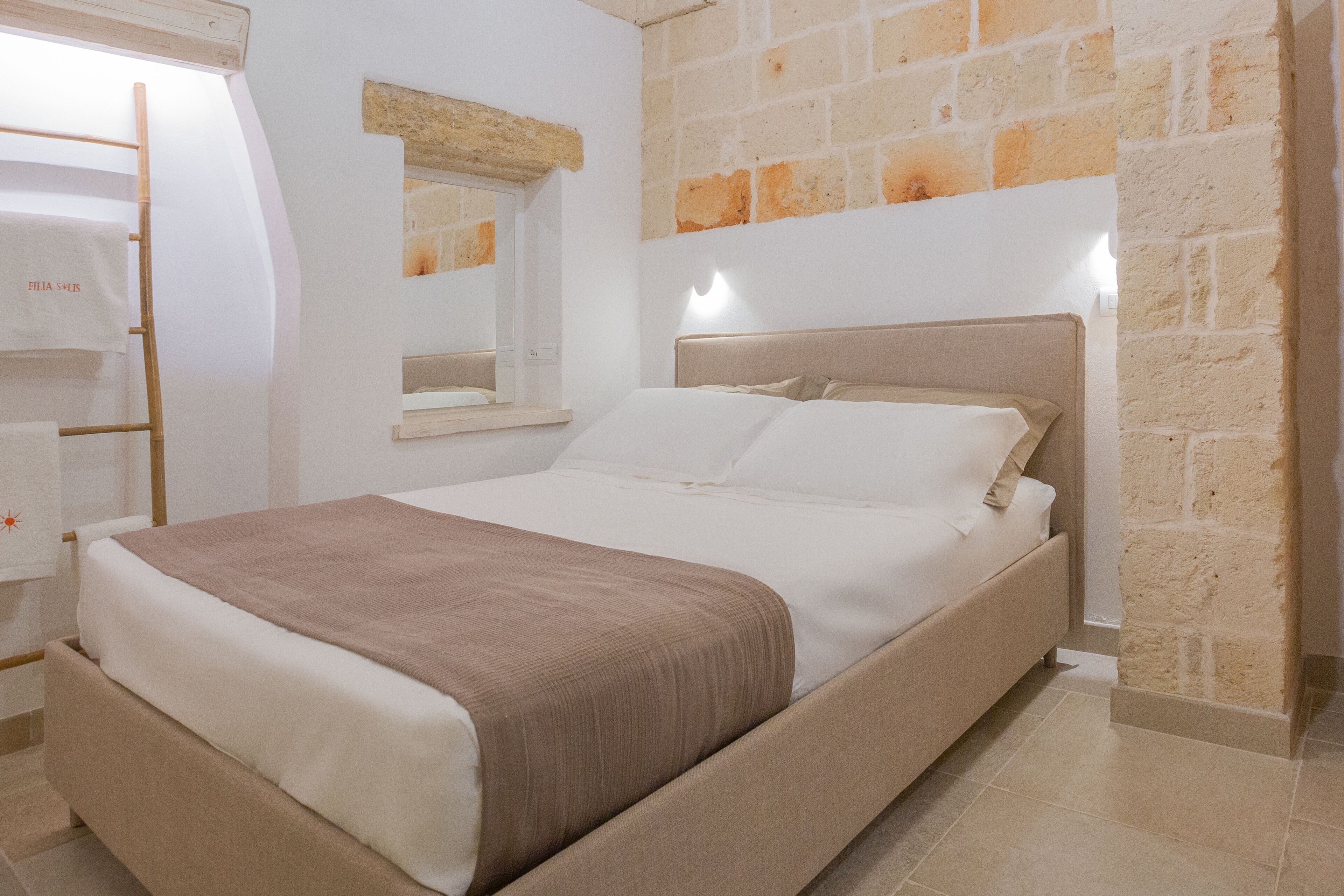 A neatly made bed with a headboard, a wooden ladder, a mirror, and a stone wall.