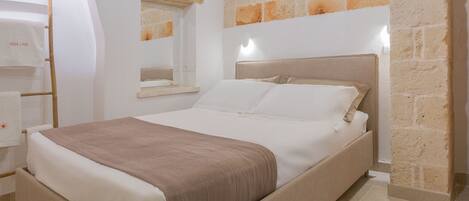 A neatly made bed with a headboard, a wooden ladder, a mirror, and a stone wall.