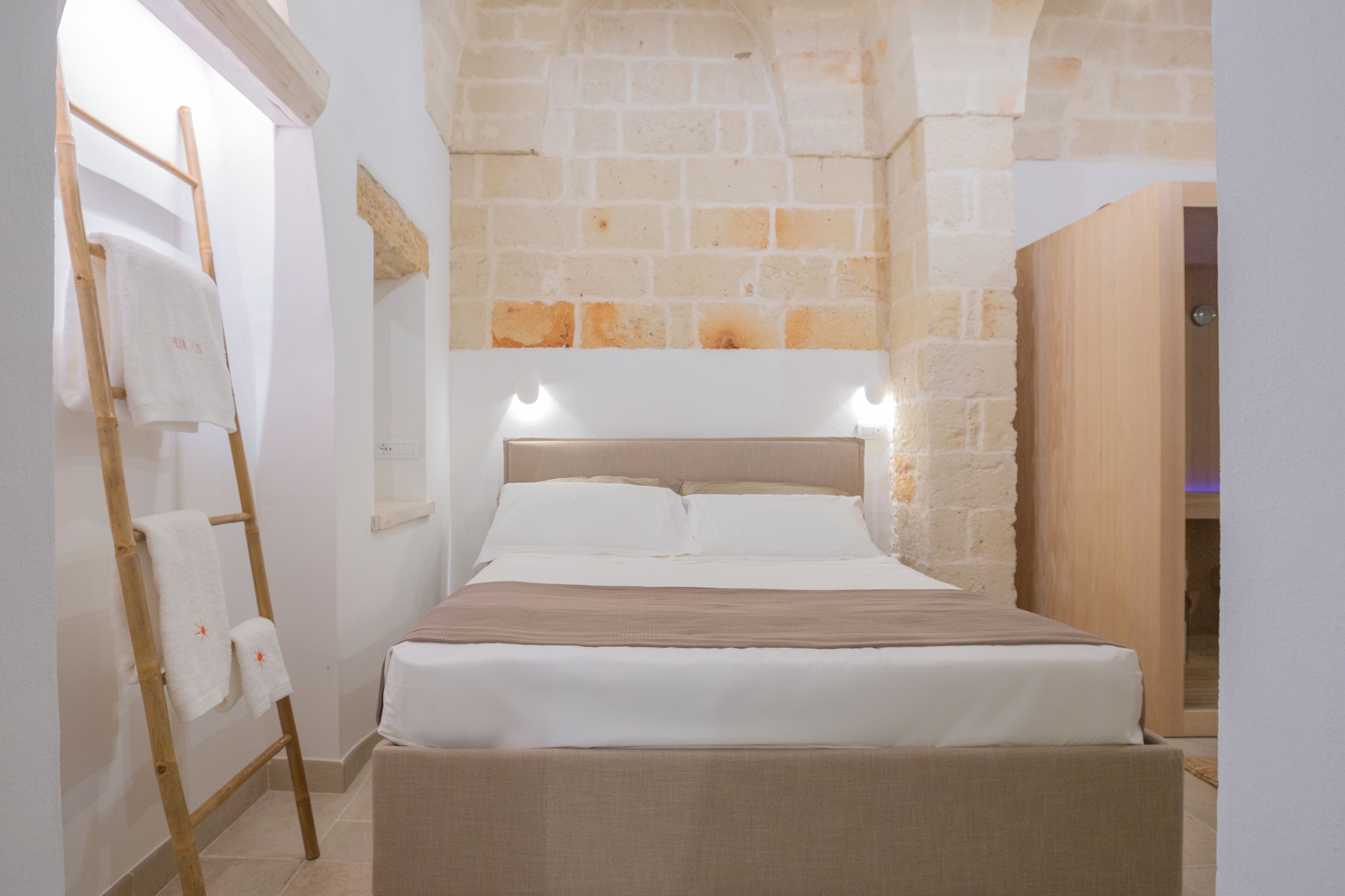 A neatly made bed in a room with stone walls and a wooden ladder with towels.