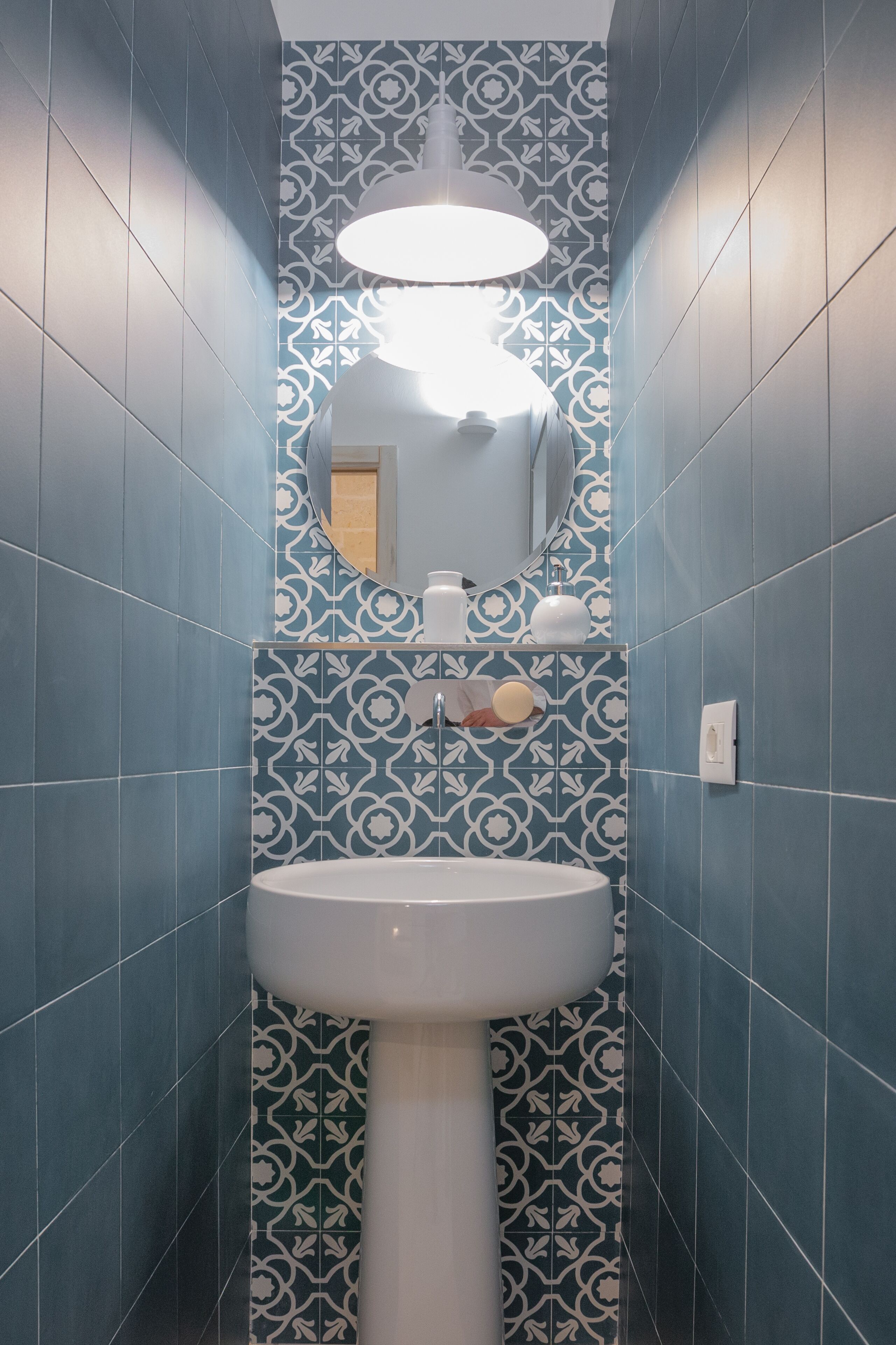Ein Badezimmer mit blauen, gemusterten Fliesen, einem weißen Standwaschbecken, einem Spiegel und einer hängenden Leuchte.