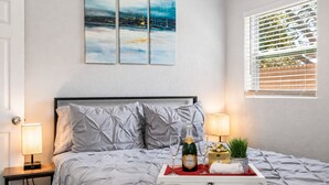 Ein Schlafzimmer mit einem Bett, zwei Nachttischlampen, einem Nachttisch, einem Fenster mit Jalousien und einem Bild an der Wand.