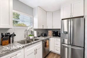 A modern kitchen with white cabinets, stainless steel appliances, a granite countertop, and a window with blinds.