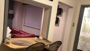 A dining area with a wooden table, two wicker chairs, a red cushion, and a window with blinds.