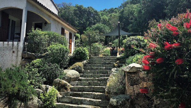 Una escalera de piedra conduce a un edificio blanco con balcón, rodeado de exuberante vegetación y flores rojas.
