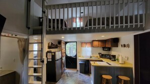 A compact kitchen with a central island, a dining area with two stools, and a staircase leading to an upper level. - Small quaint cabin with outside hot tub! sleeps 6! (Kalkaska)