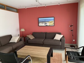 Una sala de estar con un sofá gris, una mesa de centro de madera y una silla negra. Una lámpara y una lámpara de pie están sobre el suelo.