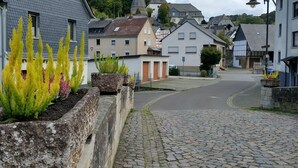 Une rue pavée dans un village aux maisons traditionnelles, avec une église et un mur de pierre orné de plantes.