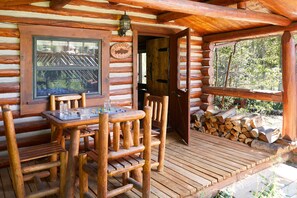 Een houten chalet met een terras, een tafel met stoelen, een raam met een welkomstbord en een deur die naar binnen leidt.