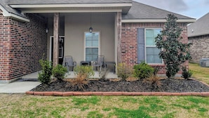A residential house with a brick exterior, a covered porch with two chairs, and a small garden area. - Cozy and Modern Home located in a New Secluded Subdivision (Lake Charles)