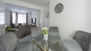 A modern living room with a glass-top dining table, grey upholstered chairs, a grey sofa with cushions, a potted plant, and a wall clock. - Fortified Three Bedroom Home (London)