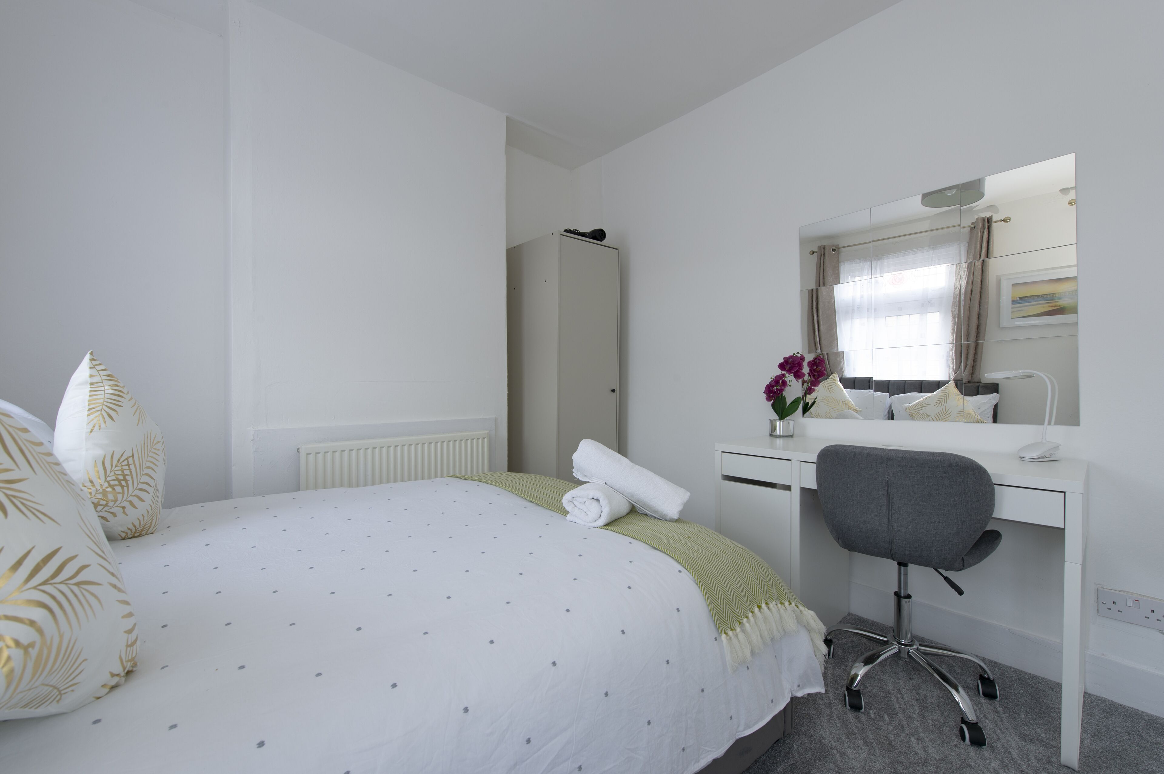 A hotel room with a bed, a desk, a chair, a radiator, a mirror, and a window with curtains.