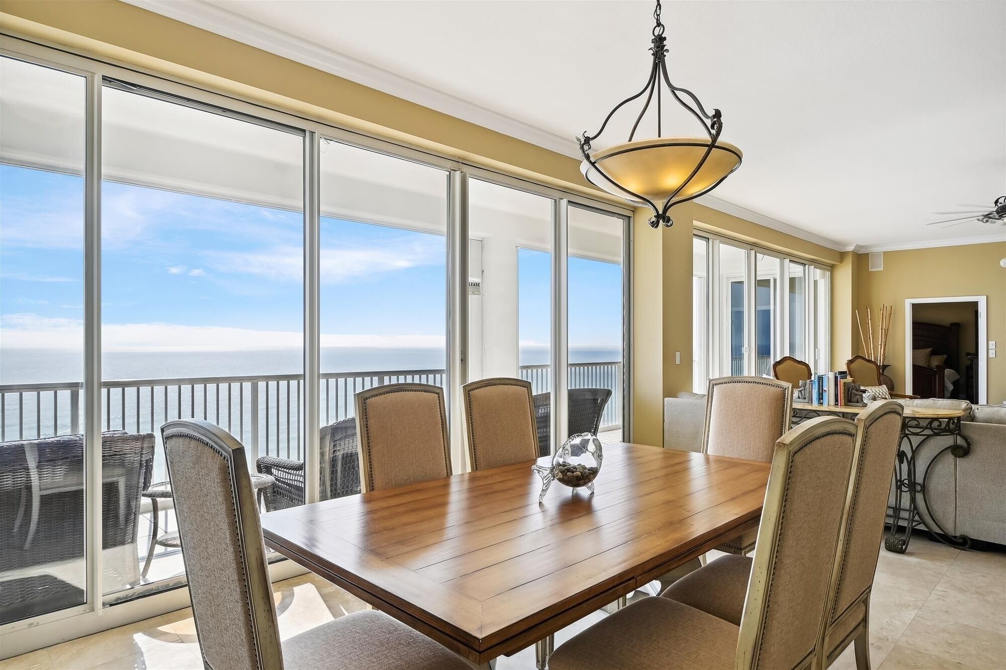 A dining area with a wooden table and chairs, large windows with a view of the ocean, and a chandelier.