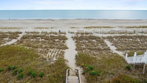 A beach with a wooden pathway leading to a white fence, sandy dunes, and a clear view of the ocean.