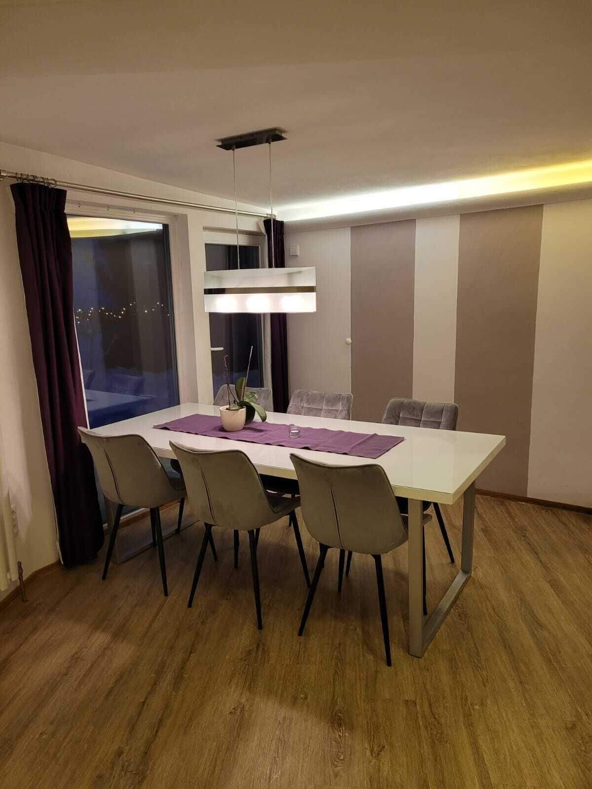 Un comedor con una mesa blanca, cuatro sillas, una lámpara colgante y vistas al exterior a través de una ventana con cortinas.