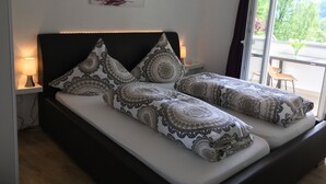Cama de casal com roupa de cama estampada, luminárias de cabeceira, uma obra de arte na parede e vista para o exterior por uma porta deslizante.