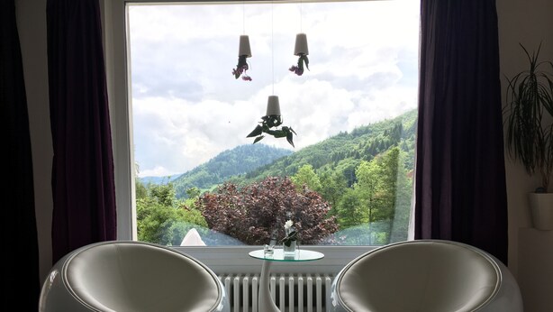 Quarto com duas cadeiras modernas, uma mesa de vidro e vista para uma paisagem verde através de uma janela com cortinas.