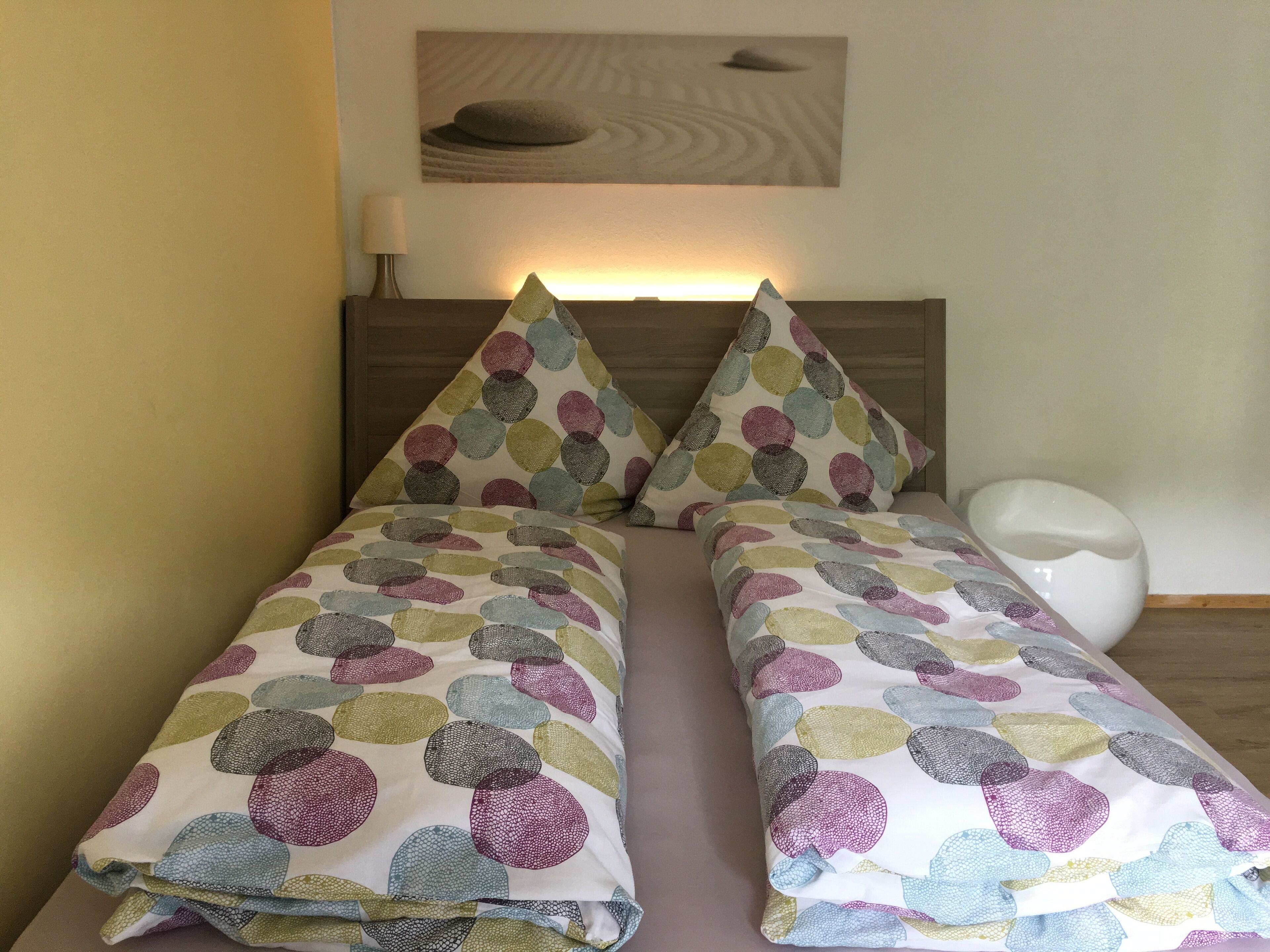 A double bed with a colorful circular pattern bedspread and matching pillows, a bedside table with a lamp, and a wall-mounted artwork above the bed.