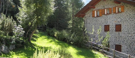 A stone building with wooden shutters, surrounded by lush greenery and a wooden fence.