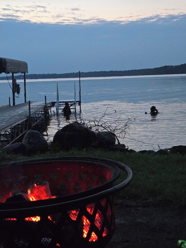 A lakeside setting with a dock, a fire pit, and a dog swimming in the water.
