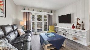 Una sala de estar con un sofá de cuero, un sillón amarillo, una mesa azul y un televisor en una pared blanca.