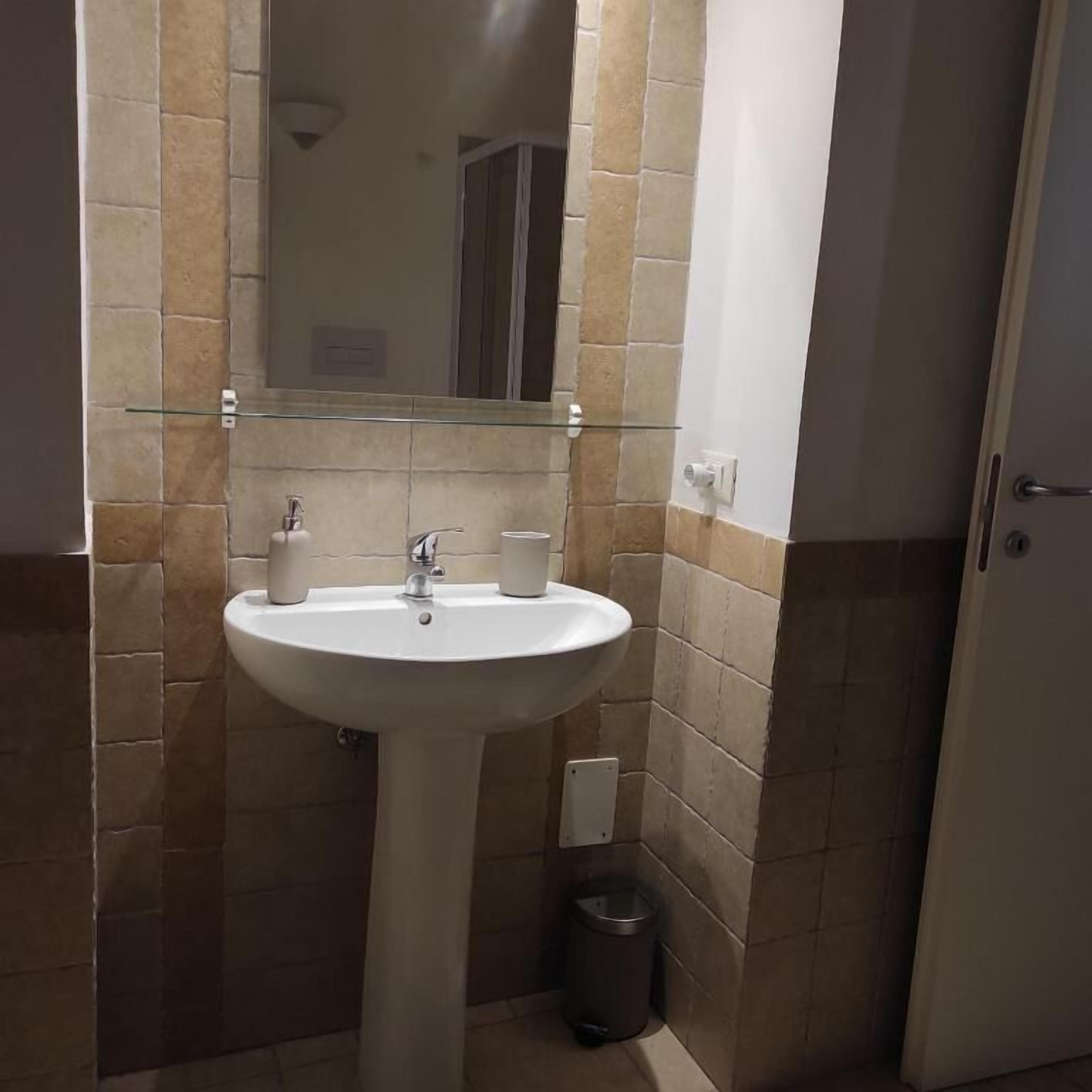 Ein Badezimmer mit einem weißen Waschtisch, einem Glasregal darüber und einem Spiegel. Ein Mülleimer befindet sich rechts neben dem Waschbecken.