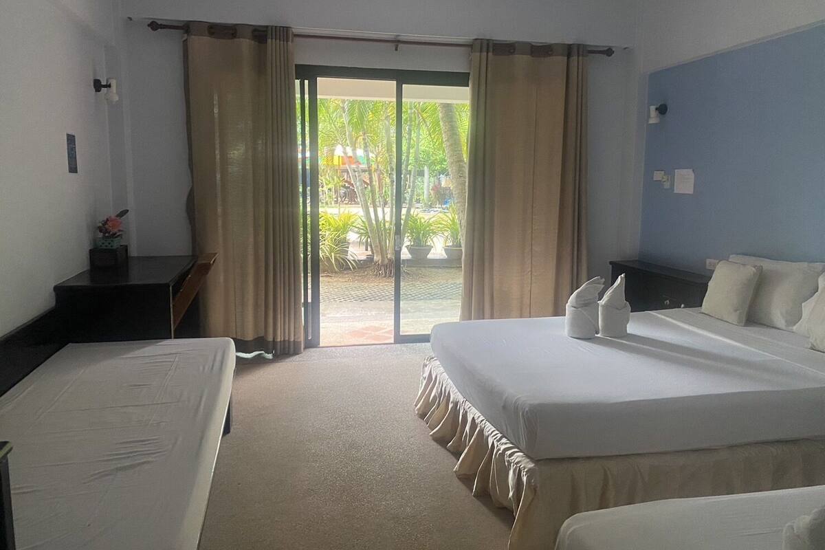 A hotel room with two beds, a desk, and a view through a glass door to an outdoor area.