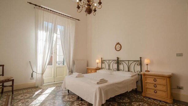 Un dormitorio con una cama, dos mesitas de noche con lámparas, una silla, un ventanal con cortinas y un suelo con diseño.