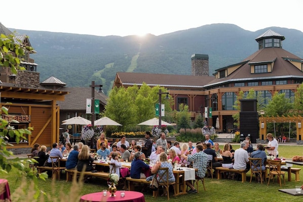 An outdoor dining area with tables and chairs, surrounded by a building with a stone facade and a mountain backdrop.