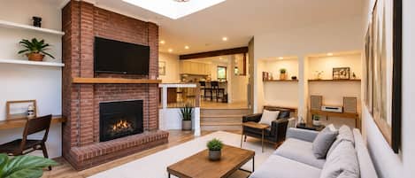 A modern living room with a fireplace, a sofa set, a coffee table, and a dining area visible in the background.