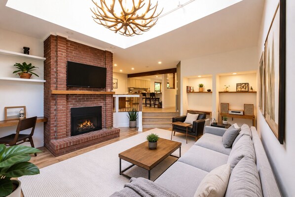 A modern living room with a fireplace, a sofa set, a coffee table, and a dining area visible in the background.