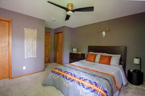 Un dormitorio con una cama, un ventilador de techo, un cráneo de ciervo montado en la pared, un tapiz de macramé y una puerta de madera.