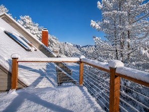 A snow-covered balcony with a wooden railing, a clear blue sky, and snow-laden trees in the background.