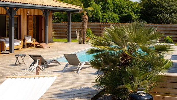 Una terraza de madera con piscina, sillones de descanso, una hamaca y una parrilla.