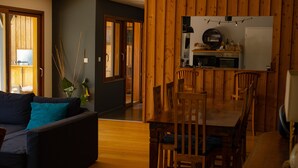 Una sala moderna con un sofá oscuro, una mesa de comedor de madera y sillas. Se ve un área de cocina al fondo con gabinetes de madera y una encimera.