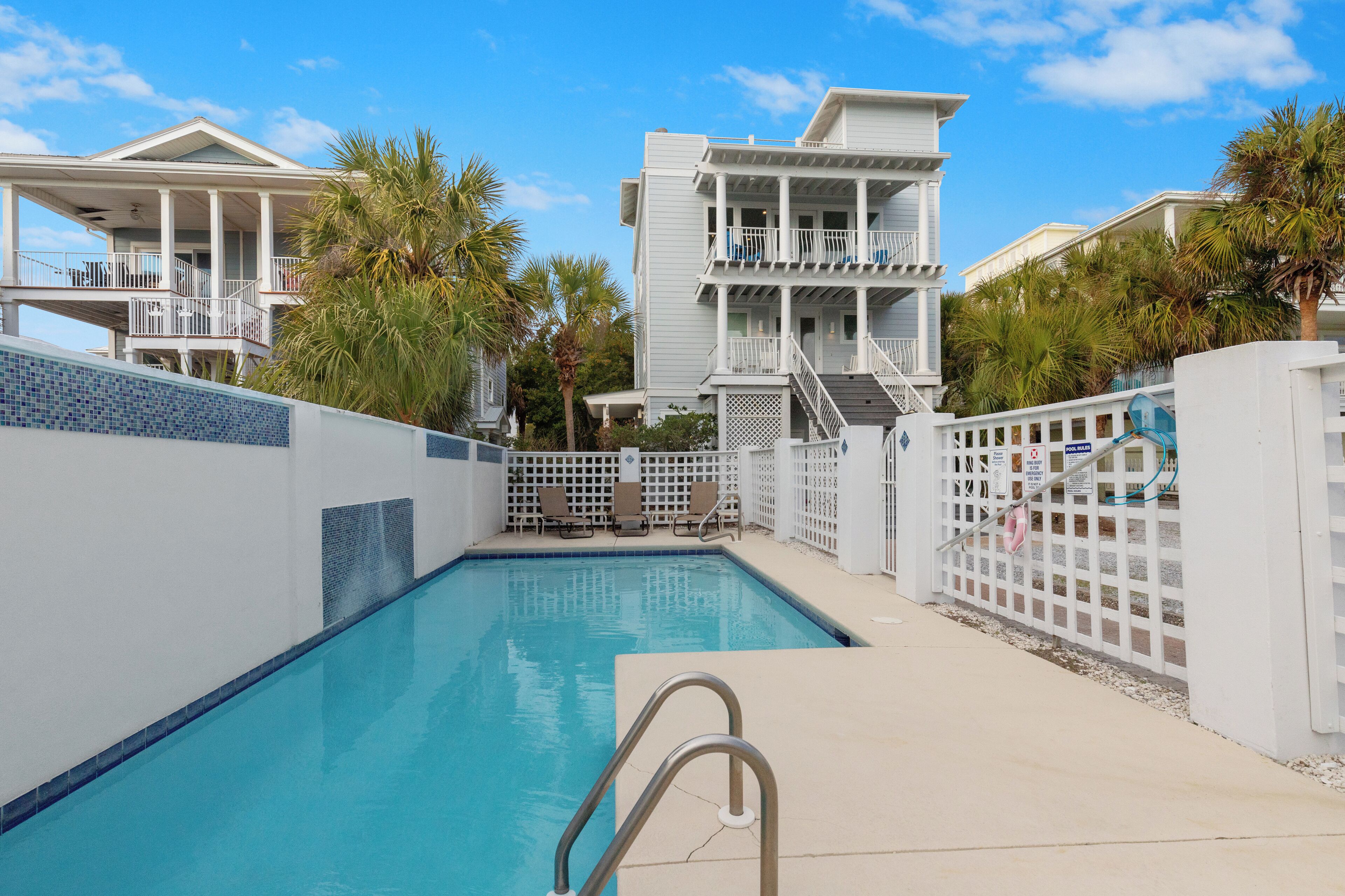 A swimming pool with a white fence, a two-story building with balconies, and palm trees.