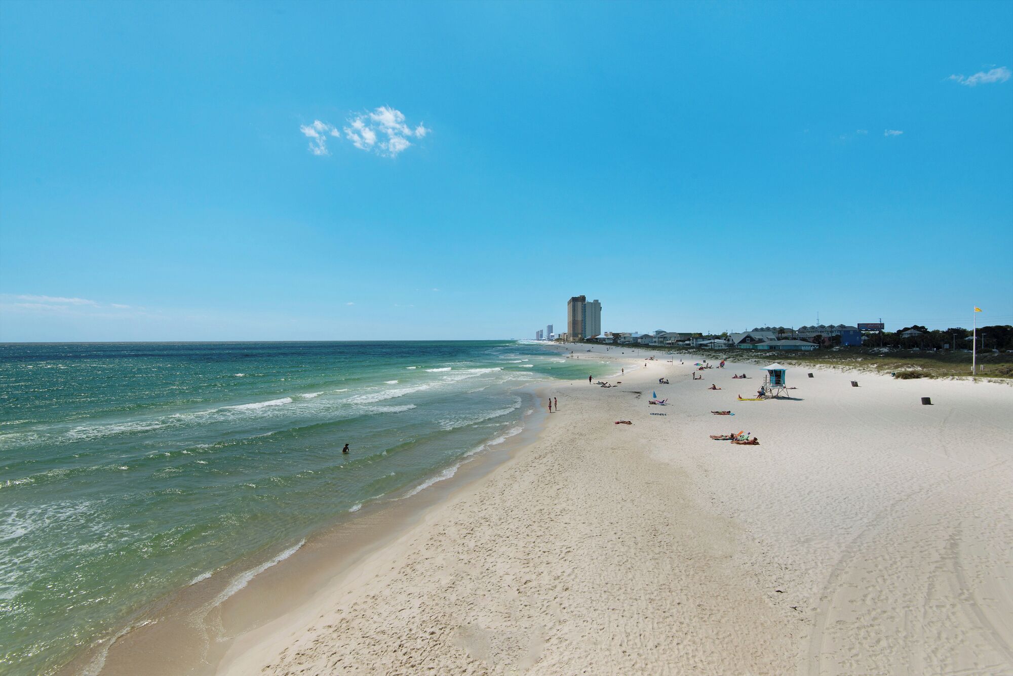 Une vaste plage de sable fin bordée d’une eau d’un bleu limpide, avec une tour de surveillance et des bâtiments en arrière-plan.