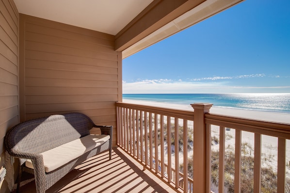 A balcony with a wicker sofa and table, overlooking a beach with sand dunes and a clear blue sky.