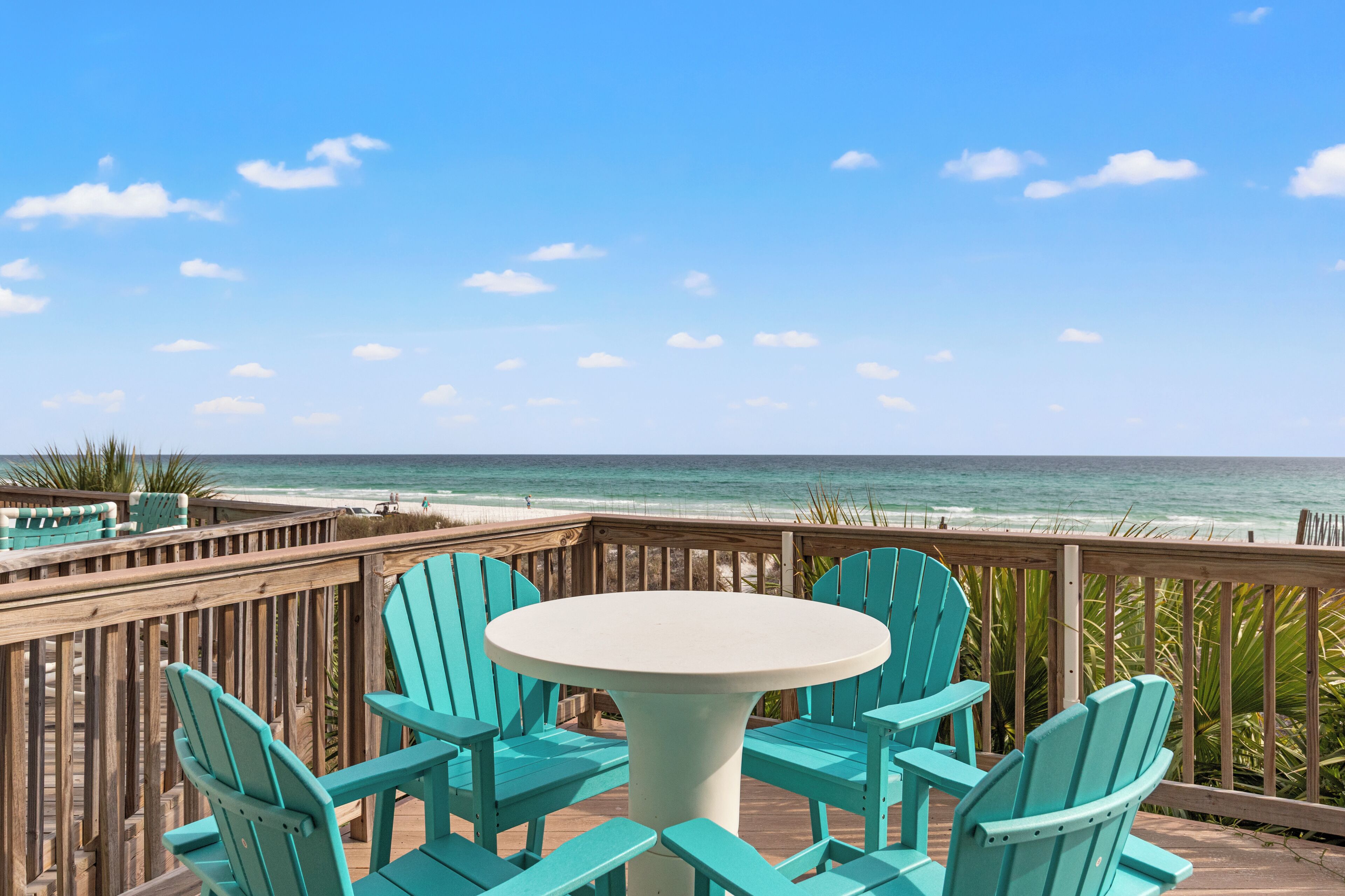 A wooden deck with turquoise chairs and a white table, overlooking a beach.