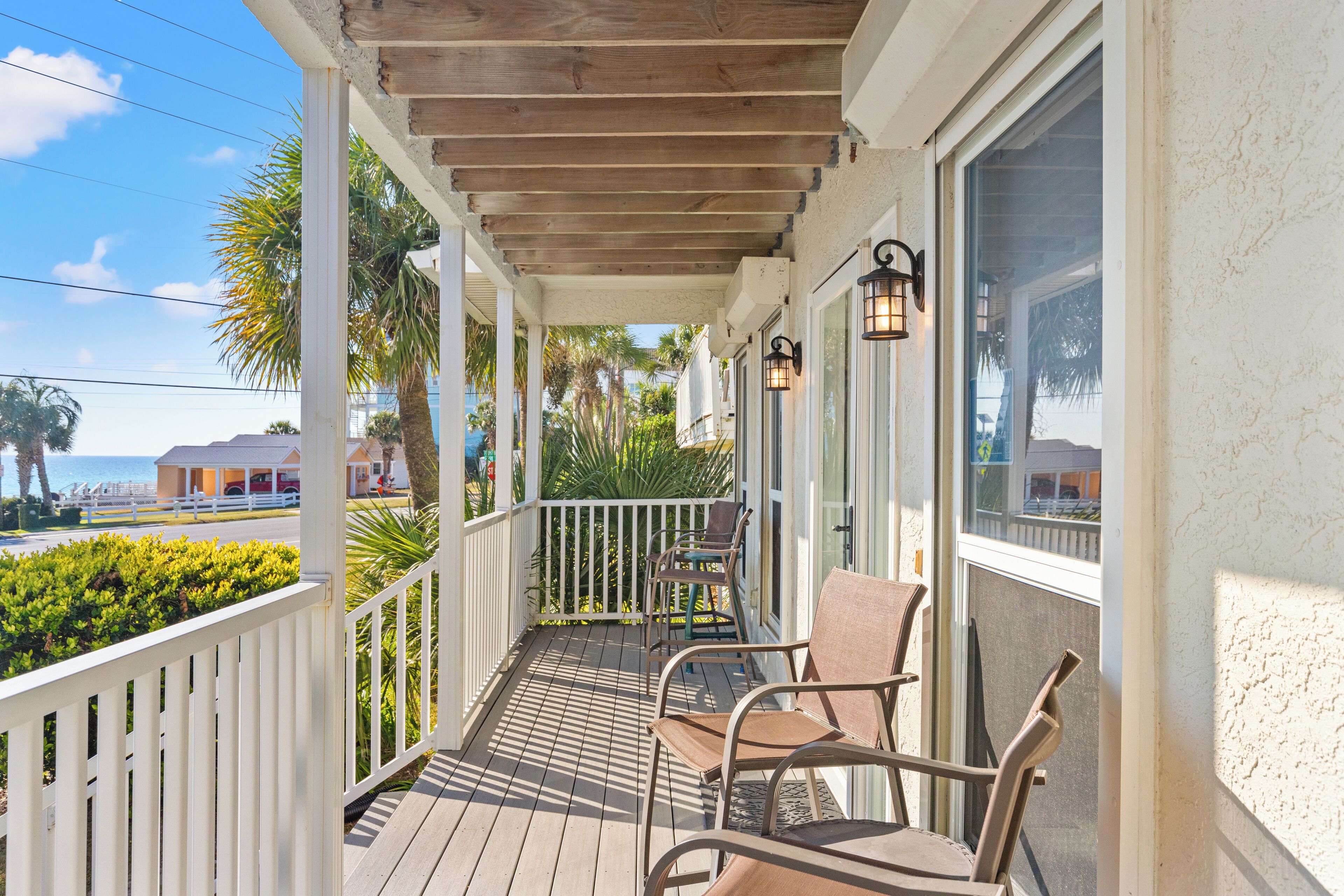 A wooden deck with chairs and a railing, overlooking a beach and palm trees.