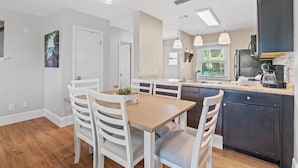 A modern kitchen with a dining area, featuring a wooden table and white chairs, a dark-colored cabinet, and pendant lighting. - 120 Palm Beach Dr A - La Casa Turquesa (Panama City Beach)