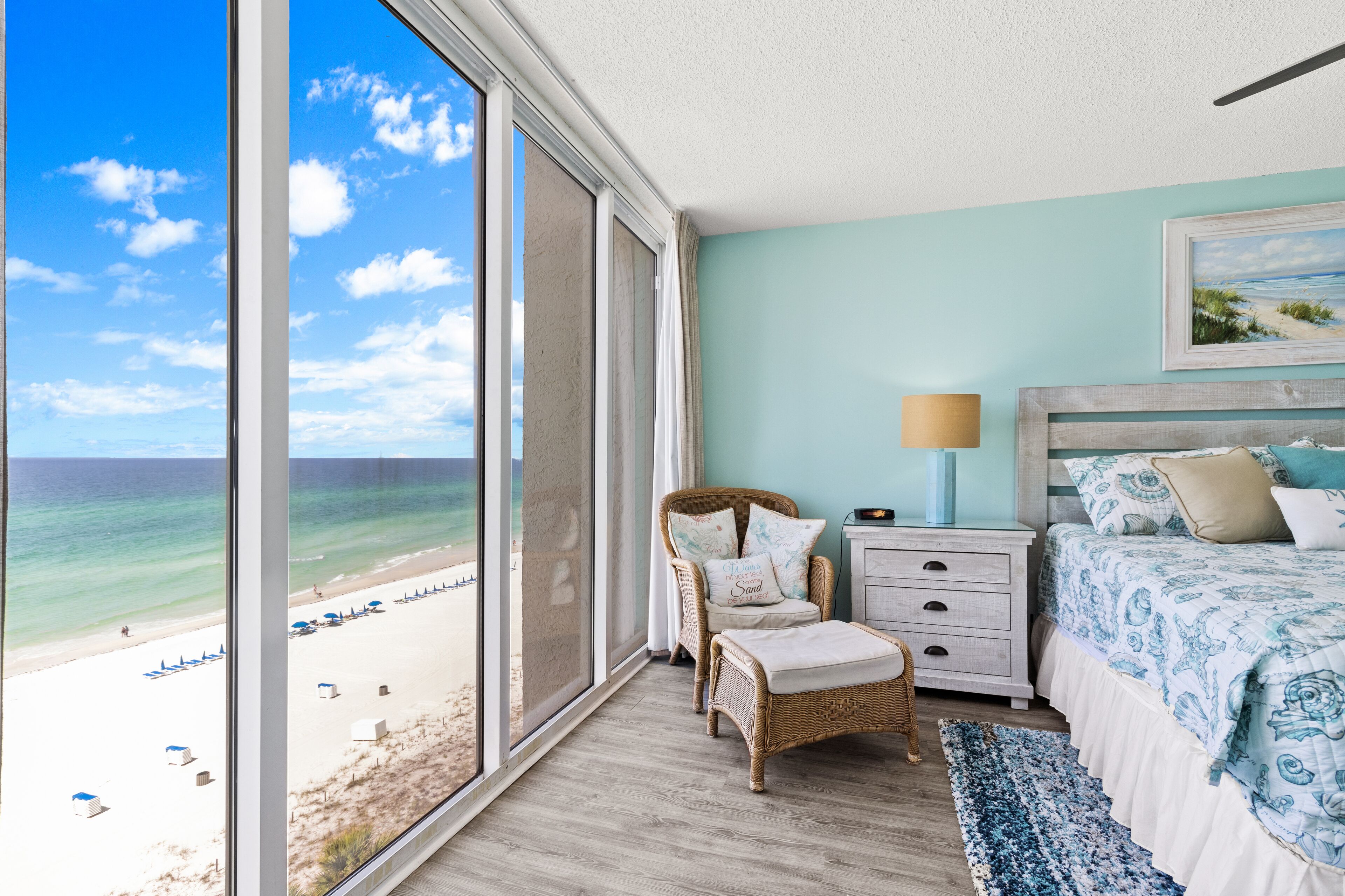 A bedroom with a large window overlooking a beach, a wicker chair, a bed with a patterned bedspread, and a nightstand with a lamp.