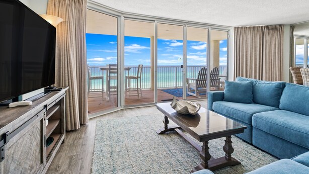 A spacious living room with a large flat-screen TV, a blue sofa, a coffee table, and a view of the ocean through large sliding glass doors. - The Perfect Escape! Long Beach Resort-8th Floor-gulf Front With Amazing Views (Panama City Beach)