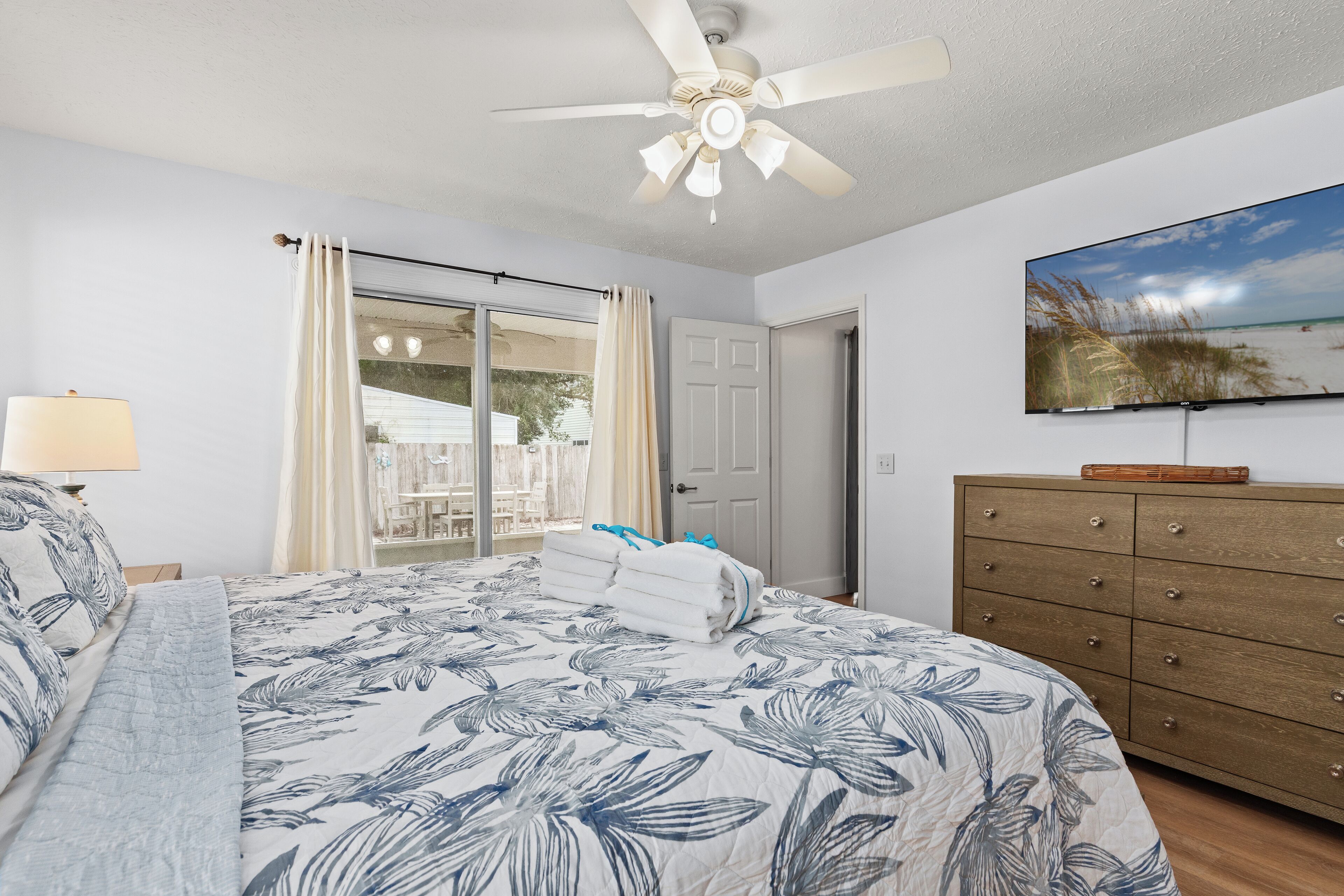 A bedroom with a bed, a ceiling fan, a TV, a wooden dresser, and a sliding glass door leading to an outdoor area.
