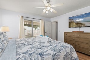 Ein Schlafzimmer mit einem Bett, einem Deckenventilator, einem Fernseher, einer Holzkommode und einer Glastür, die zu einem Außenbereich führt.