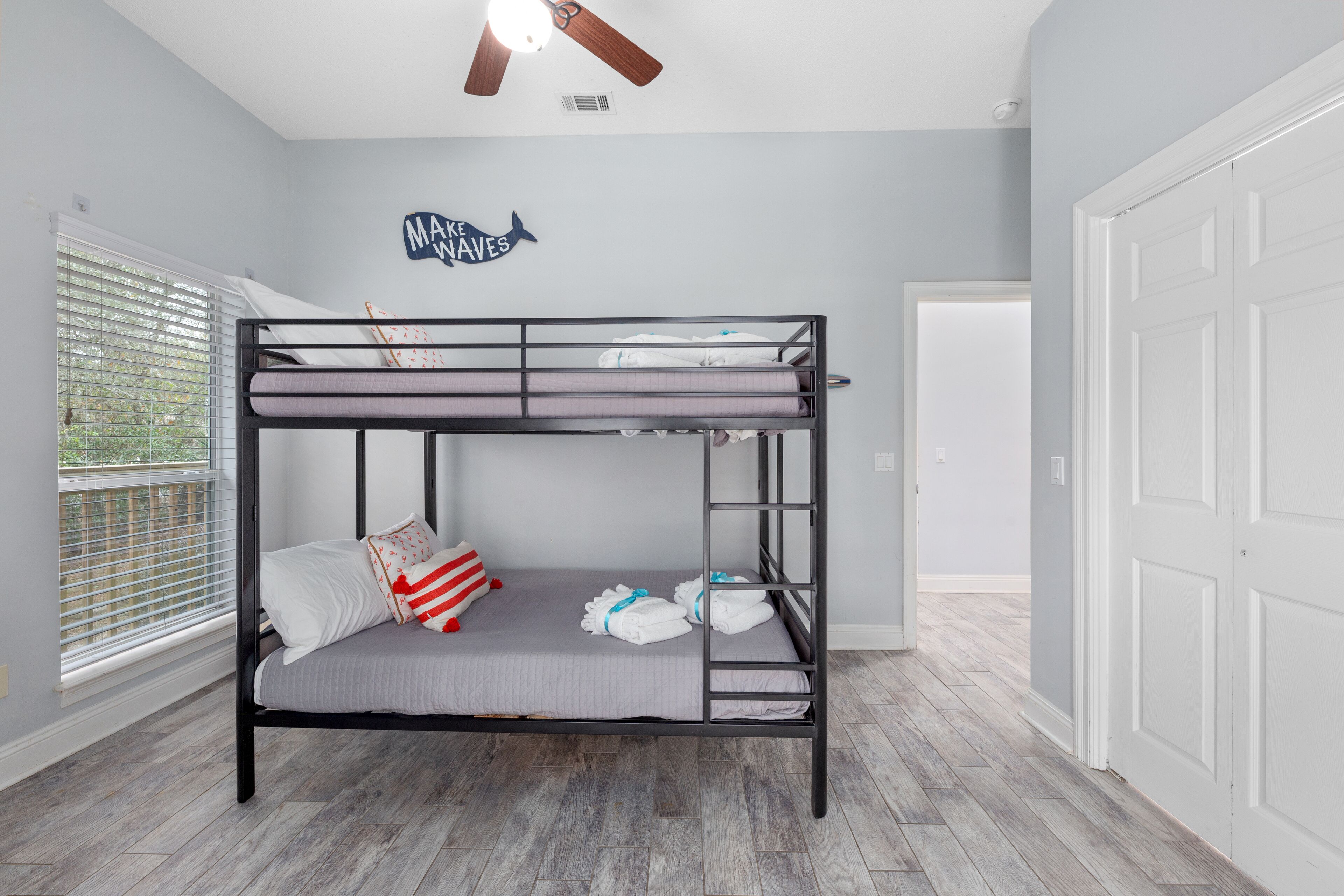 A bunk bed room with a ceiling fan, a window with blinds, a door, and a wall decoration.