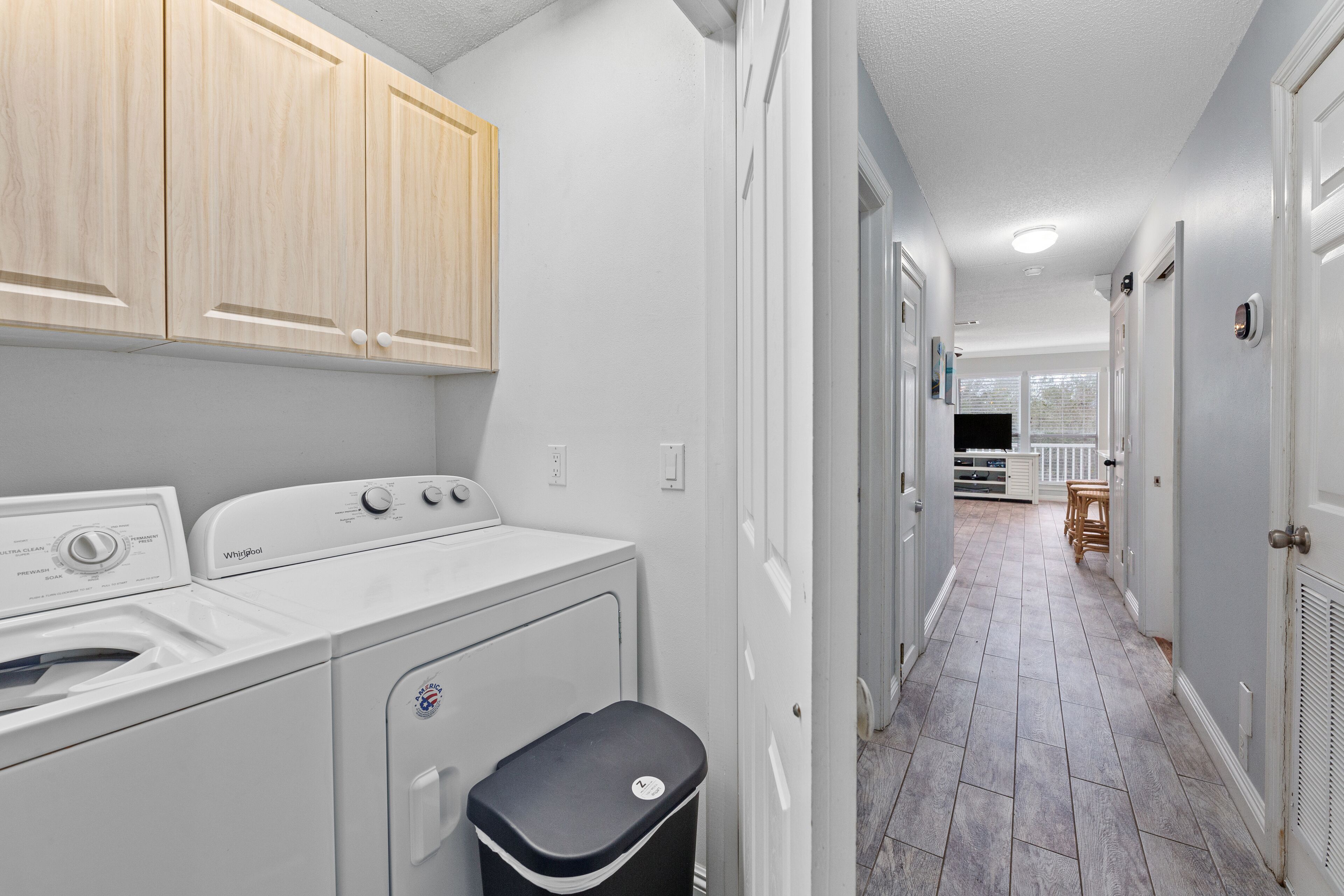 A laundry room with a washer and dryer, a trash bin, and wooden cabinets.