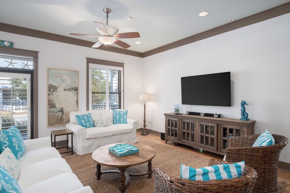 A living room with a white sofa, a round coffee table, a wicker chair, a wooden cabinet, and a flat-screen TV mounted on the wall. - Four C's Retreat - Lavish Home With Private Boardwalk Access to the Beach (Santa Rosa Beach)