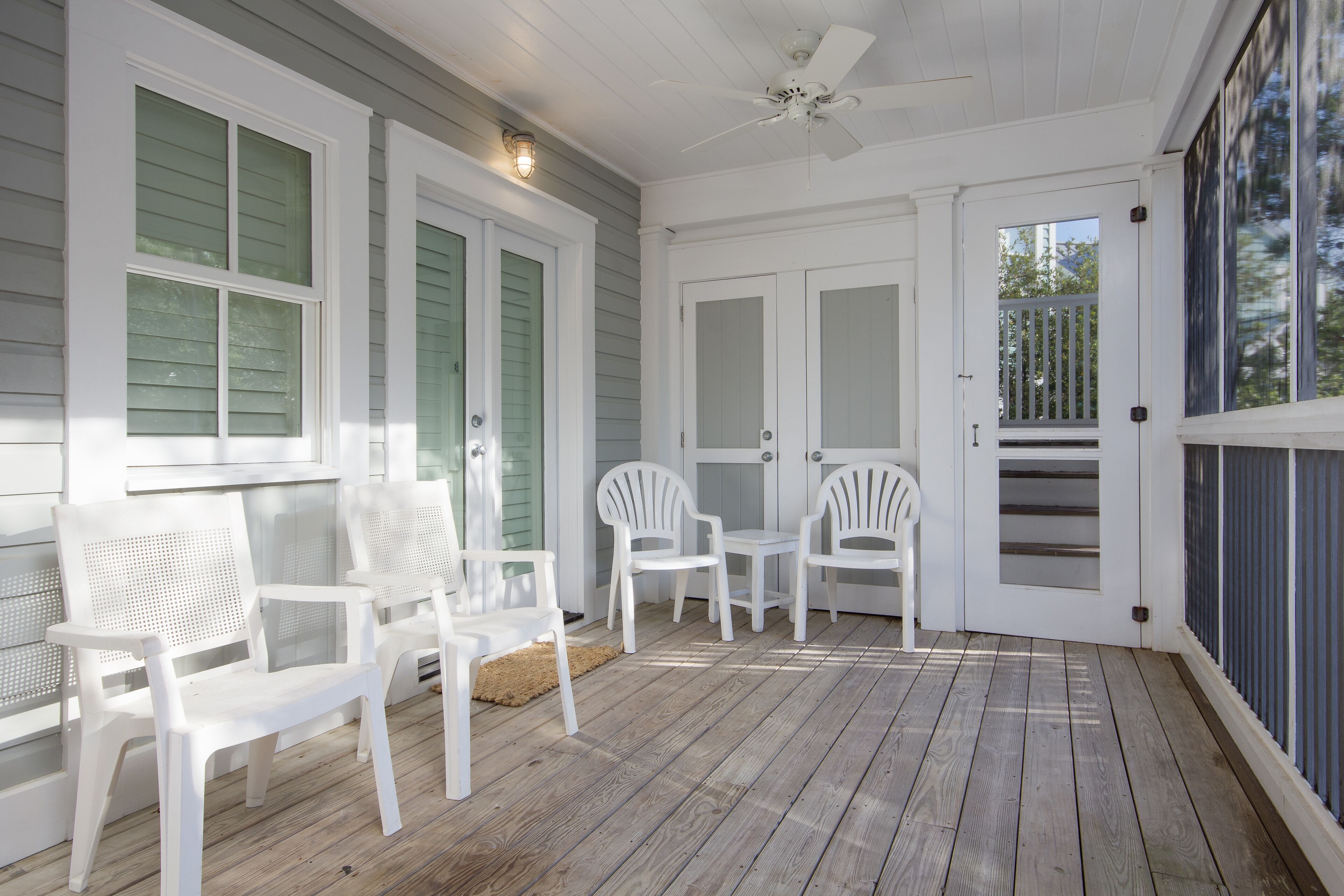 A covered porch with white chairs and a table, a ceiling fan, and a door leading to a deck.