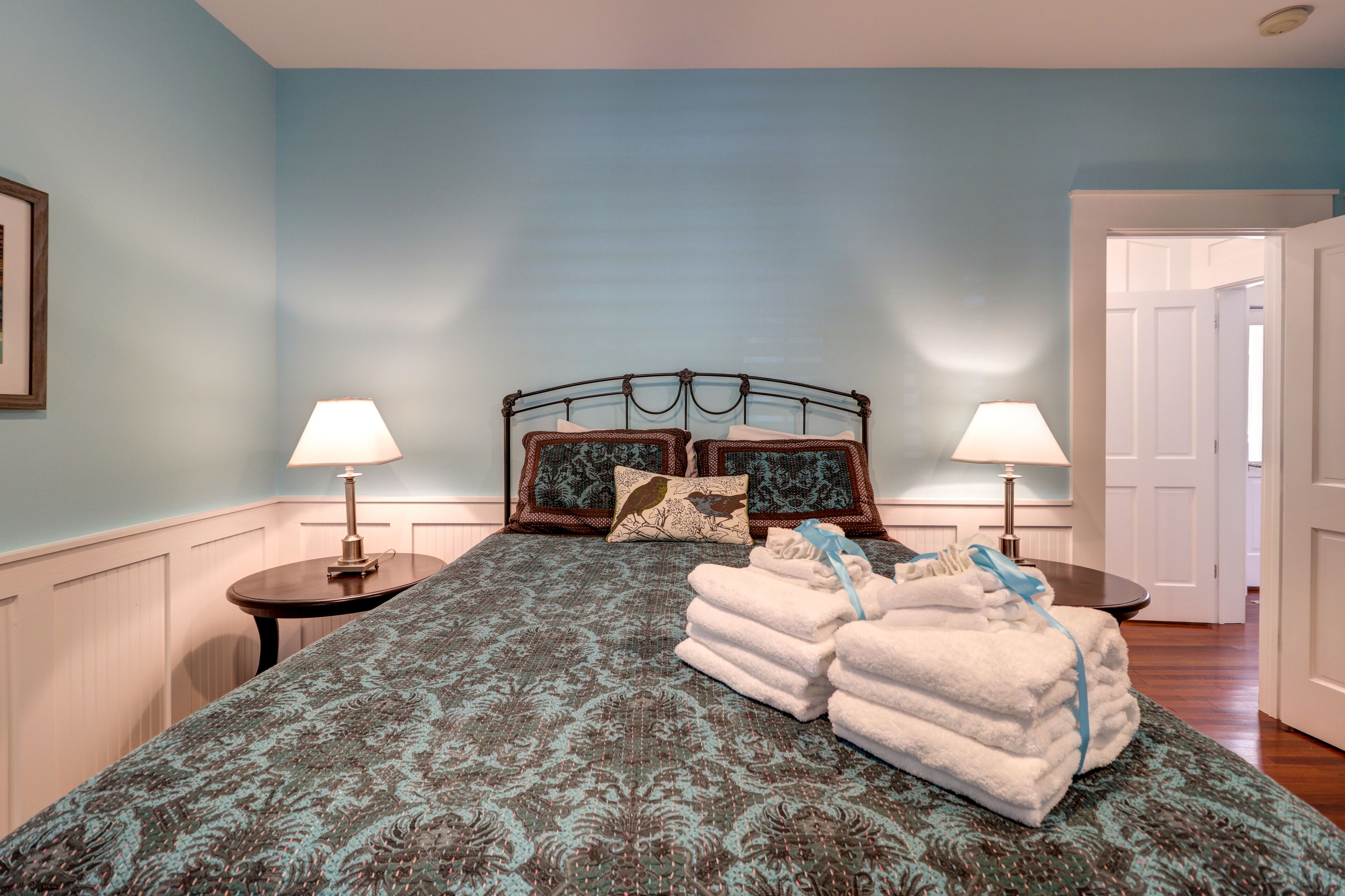 A neatly made bed with a patterned bedspread, two pillows, and a stack of folded towels. There are two bedside tables with lamps.